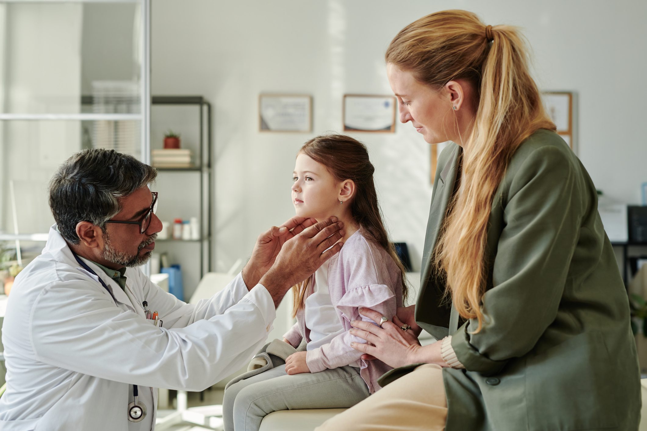 Mature male pediatrician in eyeglasses and lab coat touching thyroid of cute little girl while examining patient visiting clinics with mother